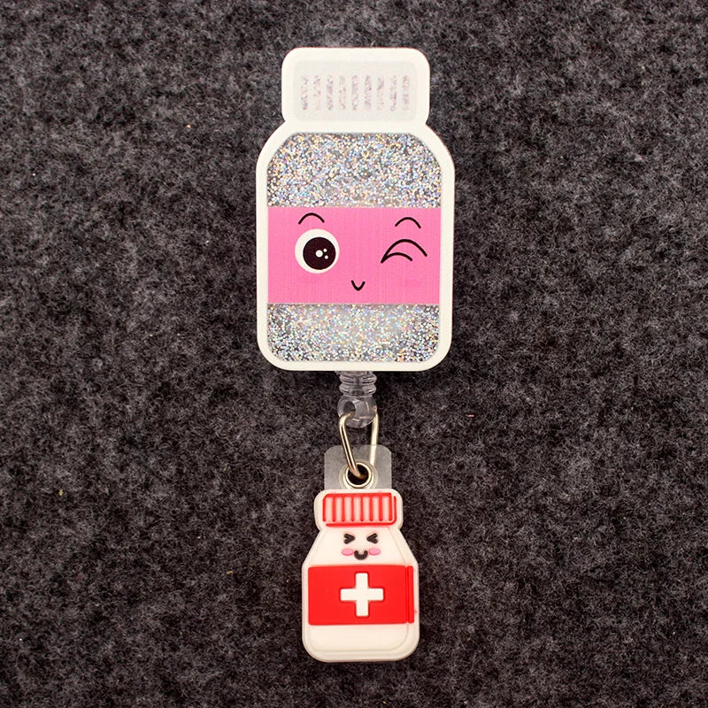 badge reel with a glittery pill bottle with a winky emoji. Under ID holder clasp, a pill bottle with a closed eye smiley emoji face